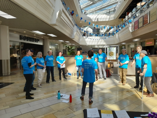 The choir standing in a semi-circle wearing blue, performing in the galleries for a mental health fair in 2022