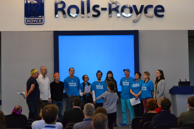 The choir standing in a huddle performing at a conference at Rolls-Royce in 2015