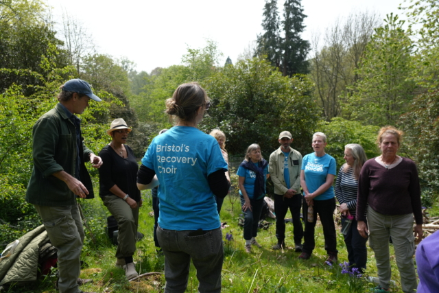 The choir standing outside in woods rehearsing for their performance at Tortworth Arboretum.