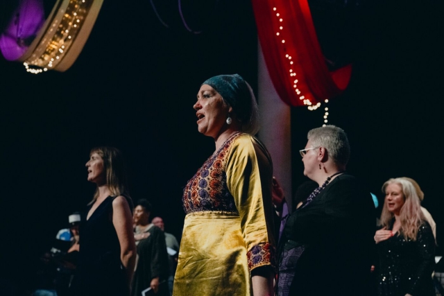 Rising Voices standing spread out on stage with their hands on their chests singing as part of the performance