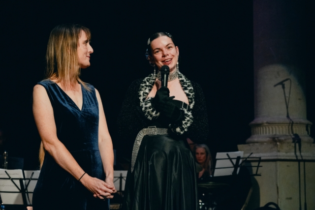 A choir member on stage introducing a song at the Creative Communities 10 year anniversary gala at Circomedia