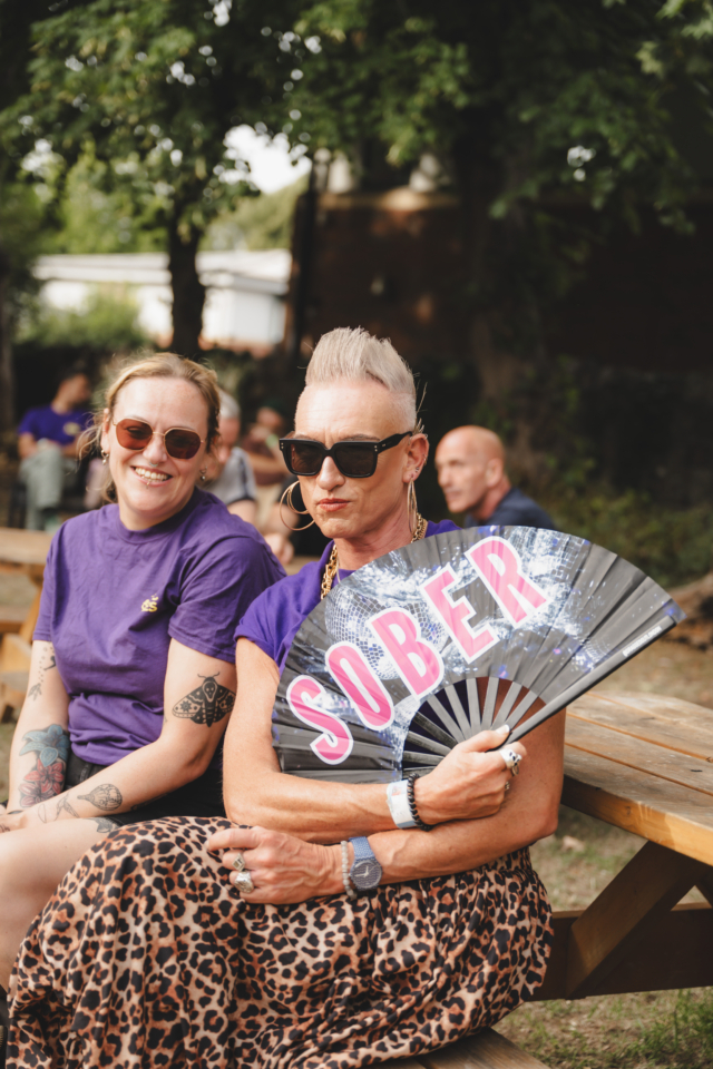 Two Sober Spaces volunteers sat together smiling for a photo holding up a fan reading #Sober