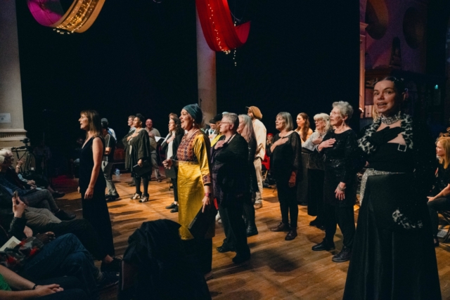 Rising Voices standing spread out on stage with their hands on their chests singing as part of the performance