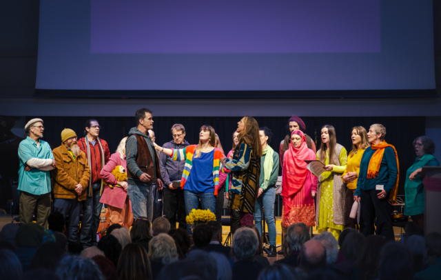 The choir standing in a line in bright colour clothes on stage performing at Maggie's memorial at St George's