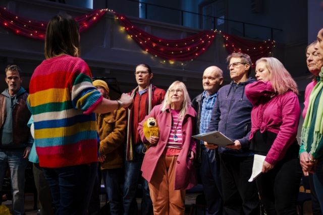 The choir standing in a line in bright colour clothes on stage at performing at Maggie Telfer's Celebration of Life at St George's